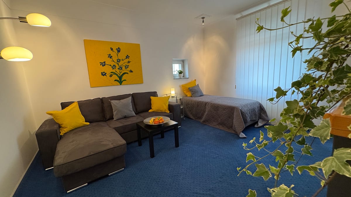 Wohnzimmer mit grauem Sofa, gelben Kissen und zusätzlichem Einzelbett